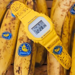A yellow digital watch rests on a bunch of spotted Chiquita bananas, with stickers visible and the time reading approximately 12:20.