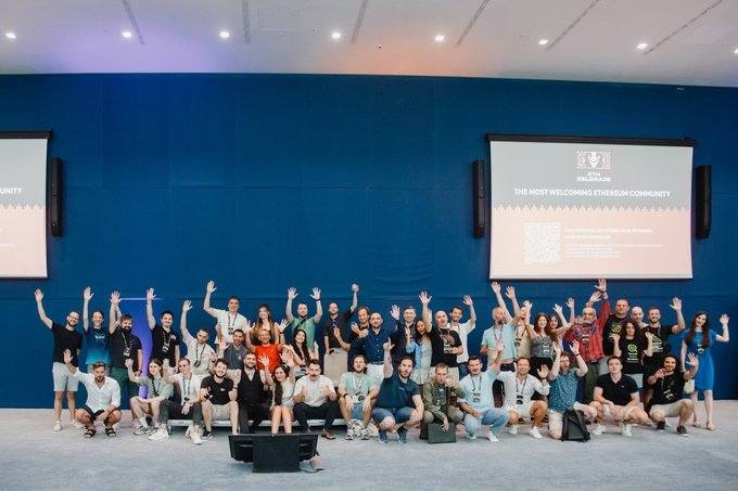 A large group photo of conference attendees posing in front of a blue wall with screens displaying 'THE MOST WELCOMING ETHERSUM COMMUNITY' and a Uniswap logo.