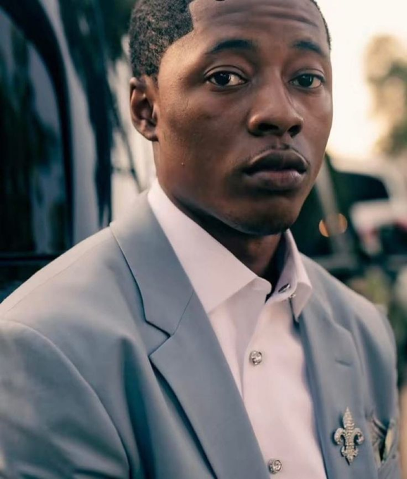 A portrait of a man in a light blue suit jacket with a fleur-de-lis lapel pin, looking directly at the camera with a serious expression.