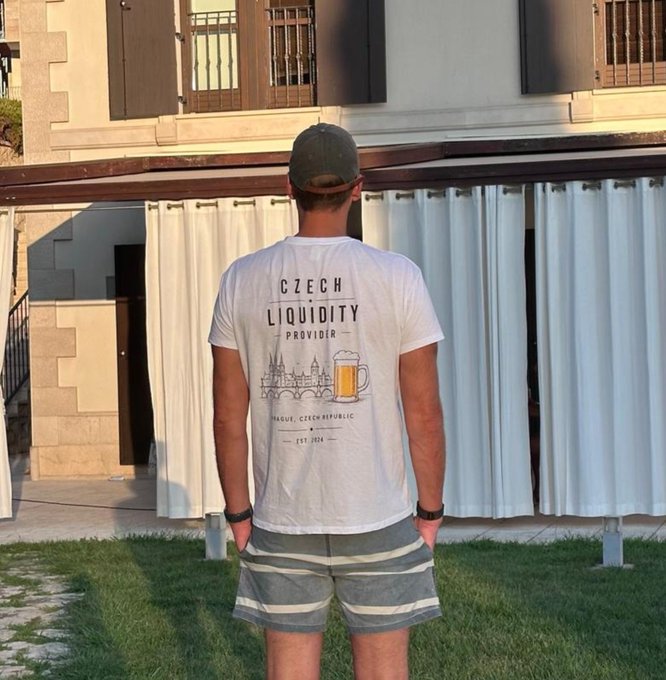 A man viewed from behind, wearing a white t-shirt that reads 'CZECH LIQUIDITY PROVIDER - PRAGUE CZECH REPUBLIC - EST 2024' with a graphic of a beer mug and city skyline, paired with striped shorts.
