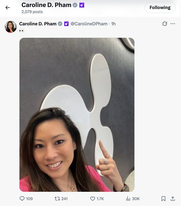 Caroline D. Pham, who has a verified account (@CarolineDPham) with 2,079 posts, is pictured smiling and pointing at a large white Ripple logo, suggesting she's at Ripple's headquarters, as noted in the accompanying post about $XRP.