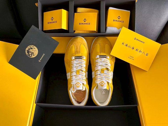 A pair of yellow sneakers with 'JOYME' branding sits in a yellow box alongside Binance-branded items, including socks, cards, and a package with Chinese text '门满山海 鞋行人客'.