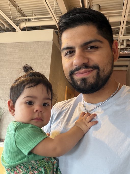 A selfie-style photo of a bearded man with a shaved head smiling warmly while holding a young child in a green top who is looking at the camera.