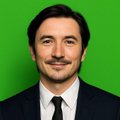 A man in a black suit and tie poses against a bright green background.