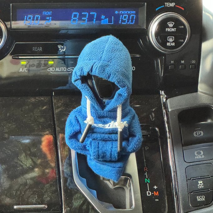 A blue knitted hooded gear shift cover with drawstrings and white text is fitted over a car's gear stick, with the dashboard display showing 8:37 and a temperature of 19.0 degrees.