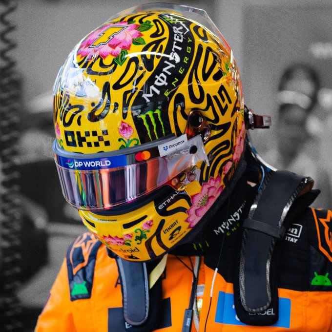 A Formula 1 driver wears a vibrant yellow helmet featuring pink floral patterns and "Monster Energy" branding, along with "DP World," "Dropbox," and "Ethara" sponsor logos, paired with a McLaren racing suit during qualifying in Singapore. The colorful tropical-themed helmet design reflects the Singapore Grand Prix's vibrant setting, complementing the post about qualifying and aiming to secure points.