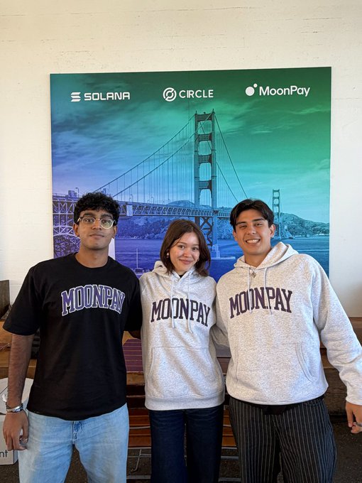 Three young people pose in front of a backdrop featuring the Golden Gate Bridge alongside logos for Solana, Circle, and MoonPay, with two of them wearing hoodies and one wearing a t-shirt, all branded with the MoonPay logo—capturing a MoonPay College merch sighting at Dev Day Cafe.