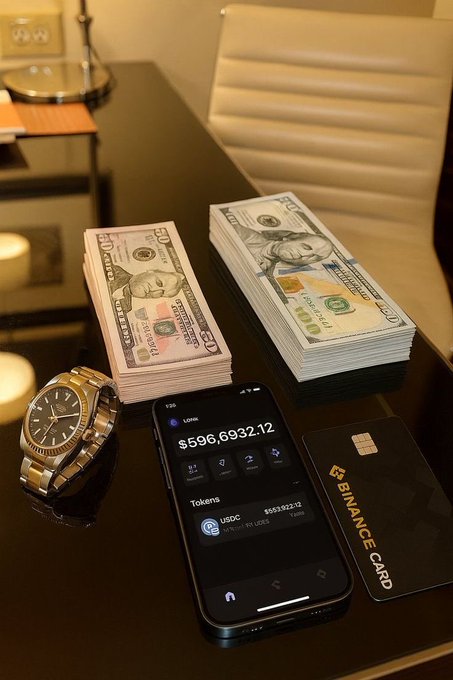 A dark wooden table displays stacks of $50 and $100 bills next to a luxury watch, a smartphone showing a balance of $596,6932.12, and a Binance Card.