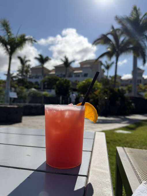 A pink/orange tropical cocktail garnished with an orange slice and a black straw, sitting on an outdoor table with palm trees and resort buildings visible in the sunny background.
