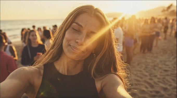 A woman takes a selfie on a beach at sunset with golden light flaring across the image and other people visible in the background.