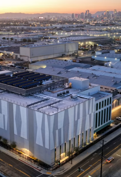 An aerial twilight view of a massive, modern industrial facility—likely an AI data center—illuminated against a backdrop of a sprawling city skyline and distant mountains, with busy roads and parking lots visible in the foreground.

The image visually reinforces the post's theme by depicting the large-scale AI infrastructure boom in America, illustrating the kind of sprawling data center investment that is now facing potential regulatory intervention in New York.