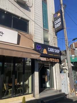 A "Moa CARD SHOP" storefront is shown, featuring prominent black signage with a yellow "M" logo and Korean text below. The image relates to the post about visiting the card shop during Consumer Crypto Day and leaving a Carp King (잉어킹) card with an MBM member, though unfortunately a 9-tier (lower quality) card had to be provided.