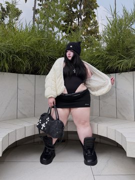 A young woman with long dark hair poses outdoors on a marble bench, wearing a white cardigan, black top, and black skirt, holding a black handbag.