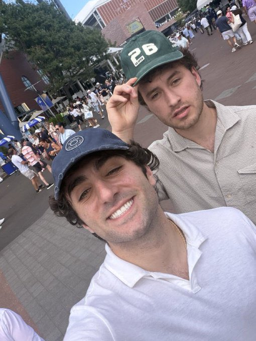 Two guys wearing caps (one with "26" and another with a circular logo) pose together at what looks like a sporting venue, smiling for a selfie with crowds in the background—capturing the smug "just got caught" energy behind the joke about insider trading on $sol and $libra.