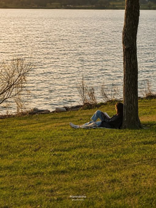 A woman in a dark jacket and blue jeans relaxes against a tree on a grassy bank beside a lake at sunset, with the watermark "Mantraksha" visible.
