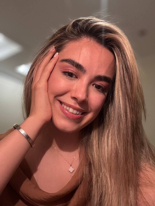 A portrait photo of a smiling young woman with long light brown hair, wearing a brown top, a gold bracelet, and a clover pendant necklace, with her hand touching her face.