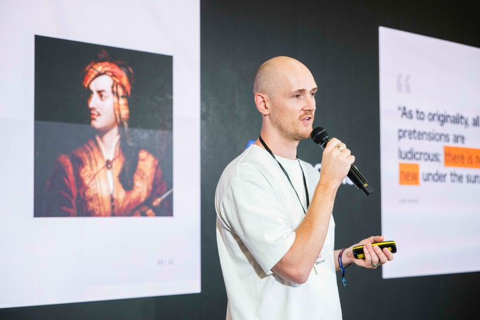 A bald man in a white t-shirt speaking into a microphone while presenting, with a classical portrait of a figure in a turban displayed on a screen behind him alongside text quoting, 'As to originality, all pretensions are ludicrous...' and 'under the sun.'