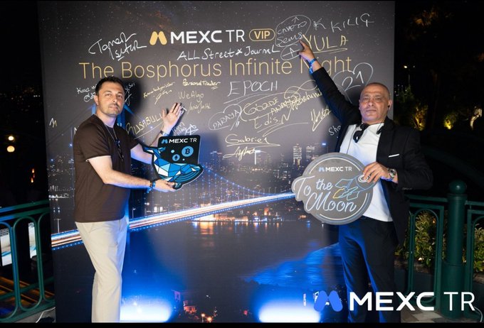The image shows two men standing next to a large black banner with white text that reads "MEXC TR". One man is holding up a sign, while the other has his arm raised high above his head. The background features a city skyline at night, which adds an urban atmosphere to the scene.