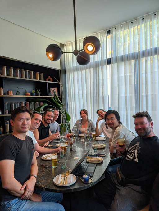 A group of eight friends gathered around a large oval dining table in a restaurant with modern decor, including a distinctive spherical chandelier and sheer curtains.