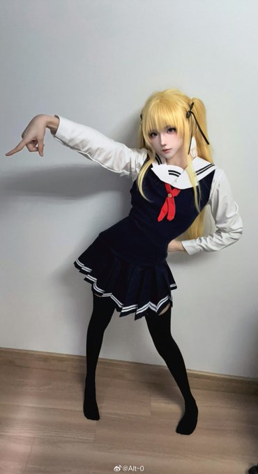 A blonde-haired cosplayer strikes a playful pose wearing a navy and white sailor-style school uniform with a red bow, paired with black tights. The watermark "@Alt-0" appears at the bottom, matching the post's username handle.