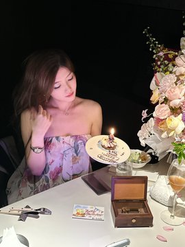 A woman celebrates at a candlelit birthday dinner with flowers, a small cake, and a wooden gift box on the table.