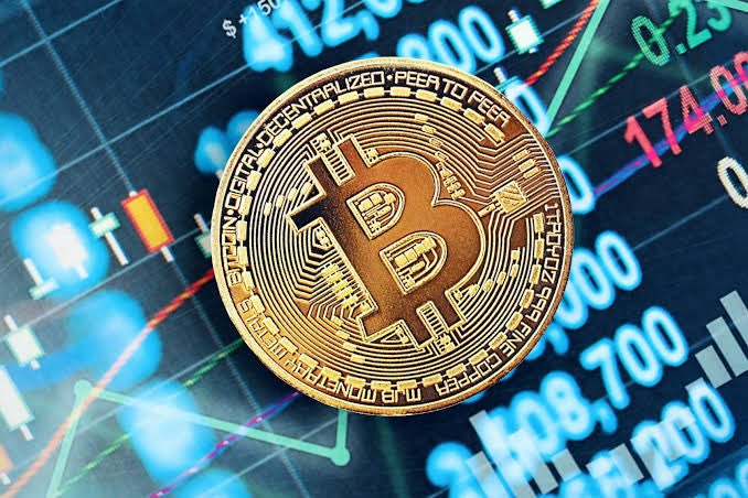 Close-up of a physical Bitcoin coin resting on a digital stock chart display showing trading numbers and candlestick patterns.