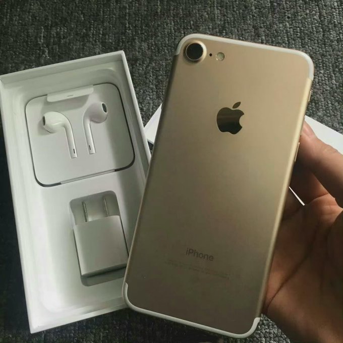 A hand holds a gold iPhone 7 next to its open box, revealing white EarPods and a power adapter inside.