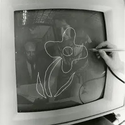 A person uses a stylus to draw a flower-like figure on the screen of an old CRT television, with the reflection of another man in a suit visible in the glass.