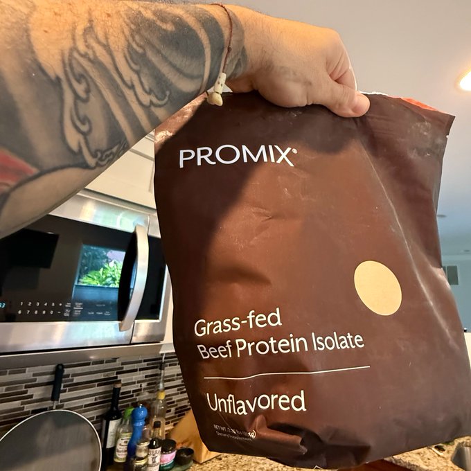 A hand holds up a brown bag of PROMIX Grass-fed Beef Protein Isolate, Unflavored, in a kitchen setting.