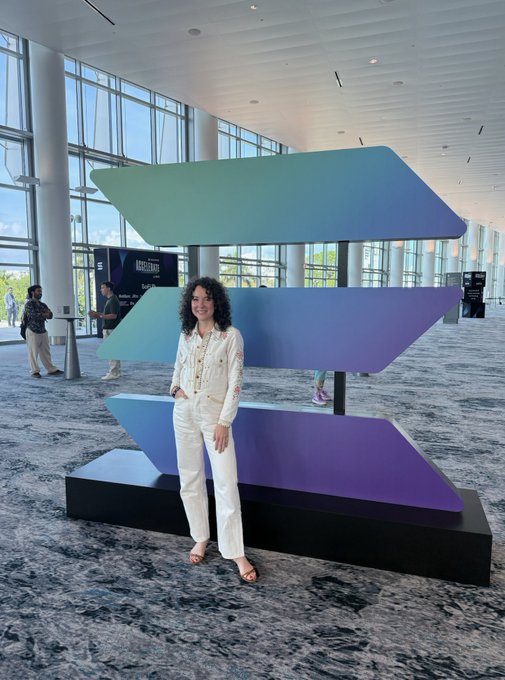 The image features a woman standing next to an abstract art piece, which appears to be a large sculpture or installation composed of multiple lines and shapes. She is wearing white pants and a white shirt, giving her a casual yet stylish appearance. The background reveals that she is in an indoor space with several people present around her. There are at least five other individuals visible in the image, some standing closer to the woman while others are further away.

The post caption reads "solana girl summer", which seems to be related to the artwork or the event taking place in this scene. The text suggests that the woman might have attended an art exhibition or a similar gathering where she encountered the abstract sculpture.