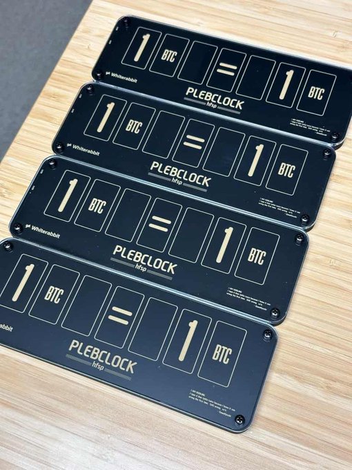 Four black signs from "Whiterabbit" rest on a wooden surface, each designed like a mechanical counter displaying "1 BTC = 1 BTC" with the text "PLEBCLOCK http".