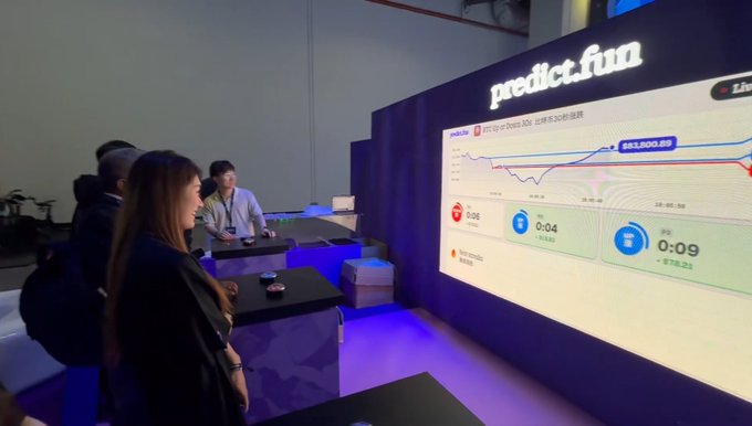 Two women view a large display screen showing cryptocurrency price charts and trading information at a 'predict.fun' booth.