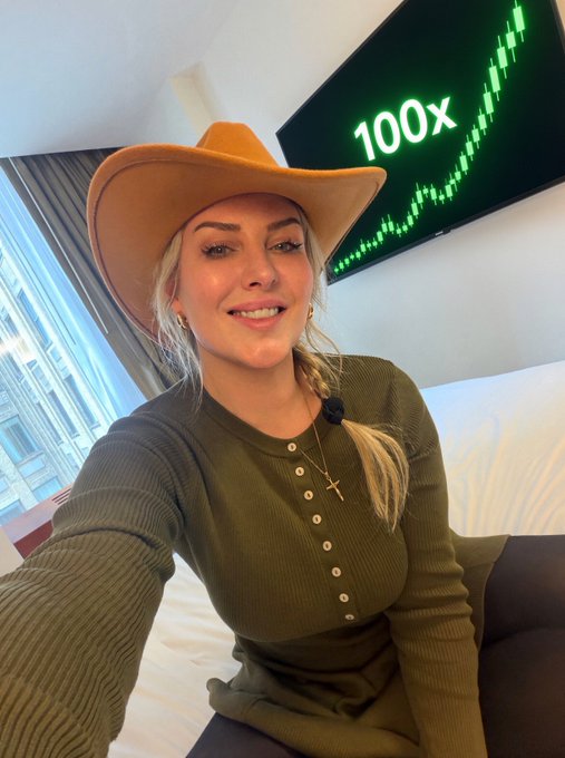 A smiling woman wearing a brown cowboy hat and an olive green ribbed henley shirt takes a selfie in a hotel room, with a TV behind her displaying '100x' next to a green stock chart graphic.
