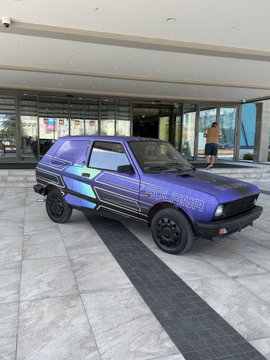 A small purple hatchback car with blue and black decals is parked outside what appears to be a modern building or dealership entrance.