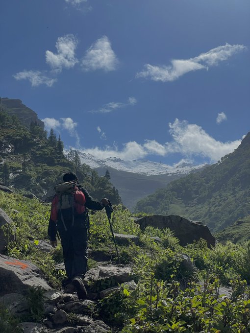 A hiker wearing a red and black backpack treks up a rocky mountain trail toward a snow-capped peak under a bright blue sky with scattered clouds.