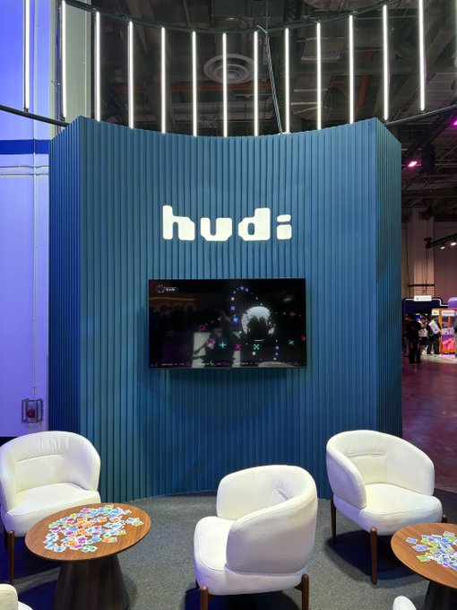 A modern exhibition booth with blue ribbed paneling displaying the "hudi" brand name in illuminated letters above a TV screen, surrounded by white armchairs and small tables with scattered stickers.