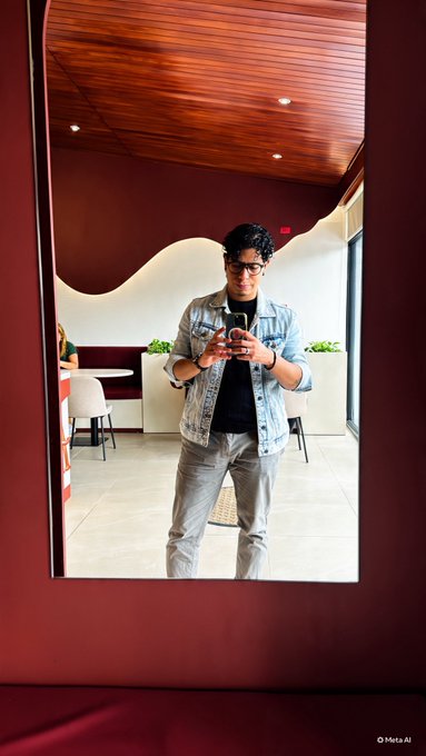 A mirror selfie taken by a man wearing glasses, a denim jacket, and gray pants, standing in a modern interior with wooden ceiling and red walls, marked 'Meta AI'.
