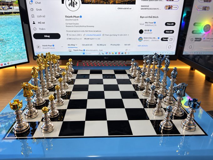 An ornate silver and gold chess set with detailed pieces arranged on a black and white board in front of a monitor showing a YouTube channel page in Vietnamese.