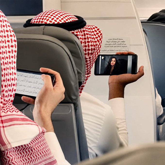 The image shows a person wearing a traditional red and white checkered Saudi keffiyeh, seated on an airplane and holding a smartphone that displays a video of a woman being interviewed, alongside an Arabic news headline that includes the phrase "سترة الحياة" (life jacket) and "تحدي" (challenge) referencing a viral trend. The post's caption "One picture but only a few will understand the message" suggests the image carries a deeper social commentary—likely critiquing how people record viral challenges and tragedies on their phones instead of offering real help, a message particularly resonant in Saudi culture.