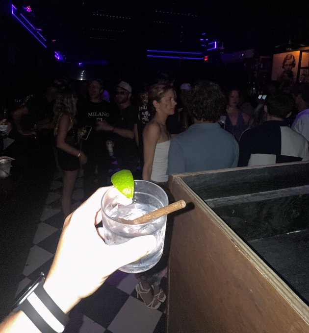 A first-person view of someone holding a clear drink garnished with a lime wedge at a dimly lit nightclub, with other patrons visible in the background under blue neon lighting.