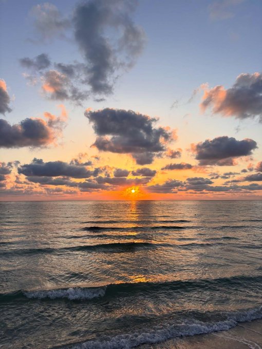A scenic sunset over the ocean with orange and pink clouds reflecting on gentle waves rolling toward a sandy beach.