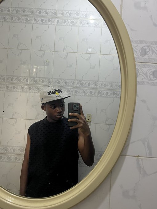 A mirror selfie of a young man wearing a black sleeveless top and a beige Wilson cap, taking the photo in a bathroom with white marble-patterned tiles.