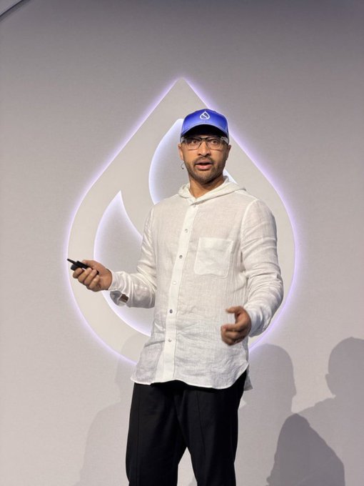 A man in a white linen shirt, blue cap, and glasses stands on stage holding a clicker in front of a large illuminated droplet logo, presenting at what appears to be a conference. The image conveys a sense of sharing market insights or "alpha" (financial intelligence), aligning with the post's invitation to discuss trading opportunities.