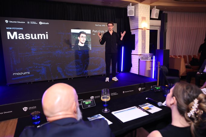 A presenter stands on stage at the "Rooftop UTXO Pitch Night" event hosted by Draper Dragon, pitching "Masumi" as displayed on the large screen behind him, with the caption noting the gathering of founders and investors focused on Cardano, Bitcoin, and the UTXO ecosystem. Judges sit in the foreground with branded materials and a glass of white wine, observing the pitch in what appears to be an upscale venue 57 floors up in Singapore.