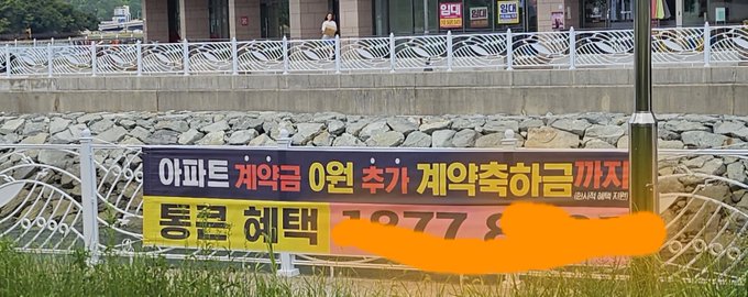 A Korean real estate sign with yellow and black sections advertises a deposit contract, featuring text like '아파트 계약금 0원 추가 계약축하금' and '통큰 혜택' along with a partially orange-redacted phone number, set against a rocky embankment.