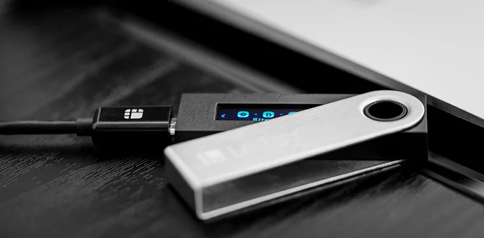 A close-up photograph shows a USB Ledger hardware wallet connected by cable to a laptop, with its small screen displaying icons.