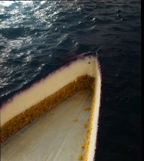 A bizarre perspective shot looking down at the inside of a large triangular object that has been cut open, revealing a pale cake-like interior with a brown crumb filling, floating in the middle of the ocean.