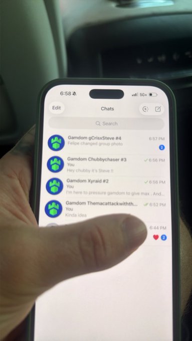 A hand holds a phone displaying the Gamdom chat app, showing active group chats named "Gamdom gCrIsxSteve #4," "Gamdom Chubbychaser #3," "Gamdom Xyraid #2," and "Gamdom Themacattackwithth..." with messages from "Steve" confirming his involvement in VIP hosting under the code "steve" for the top 25 players this month.
