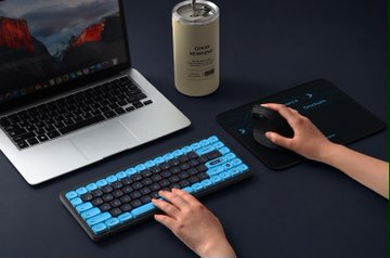 A person types on a mechanical keyboard with blue backlighting and uses a mouse on a black pad, with a laptop and a tumbler cup nearby.