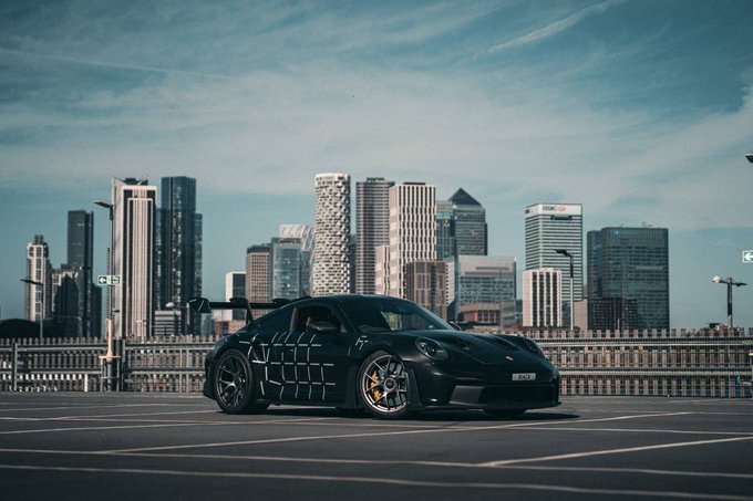 A sleek black Porsche GT3 RS with custom geometric side graphics sits in an empty parking lot against the backdrop of Canary Wharf's skyline, with a visible license plate reading "GT3 RS." The image ties into the post by showcasing one half of the giveaway prize—the custom sports car—positioned to evoke exclusivity and aspiration, reinforcing the post's promise of a $1 million prize plus this rare vehicle for trading perps on the @joinfrontier platform.